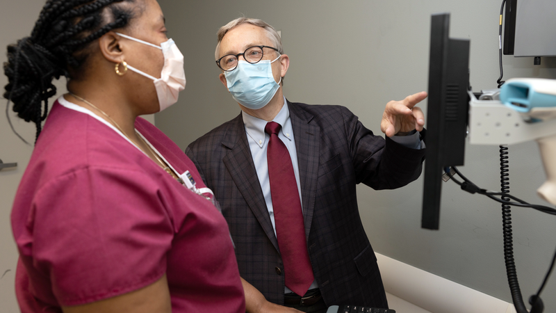 Scott Rowley points to a computer screen as he explains to a MedStar nursing associate, wearing burgundy scrubs, in a clinical setting. Both people are wearing masks.