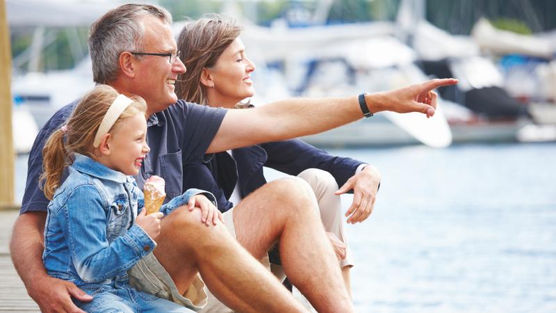 A family sits together on a pier in a marina. The little girl is eating an ice cream cone.