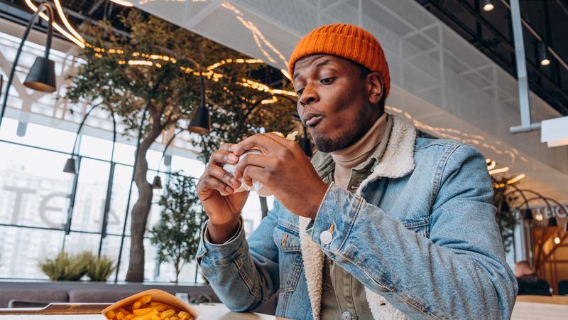 African man in an orange hat and denim jacket eats a delicious burger and French fries in a shopping mall on a food court, greasy and unhealthy fast food