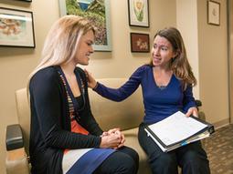 A female doctor rests her hand on her female patient's shoulder as they sit together and talk in the office waiting room. 