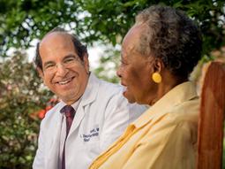 Dr. David Shocket, gastroenterologist, sits and talks with a female patient. Both are sitting together on a bench in a garden.