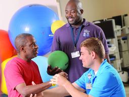 2 physical therapists work with a patient at MedStar Good Samaritan Hospital's rehabilitation center. The patient is seated and there are colorful therapy balls in the background.