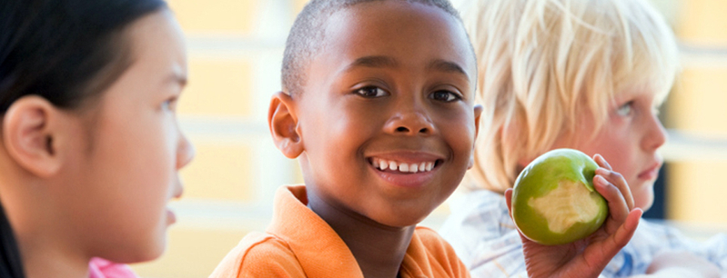 A young boy smiles as he eats an apple in a cafeteria at school.