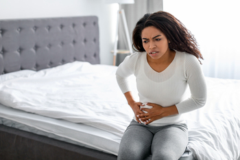 A young woman sits on the edge of her bed and winces in pain.