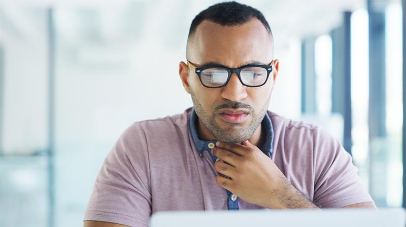 A man touches his throat and grimaces, while looking at a laptop screen.