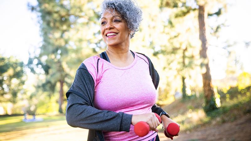 A mature african american woman walks outdoors in a park while holding hand weights.