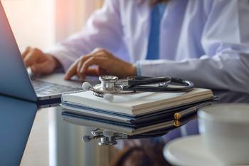 Female doctor in white lab coat typing on laptop computer with notebook and medical stethoscope on the desk in an office.