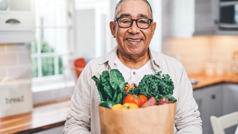 A mature man holds a bag of groceries while standing in his kitchen.