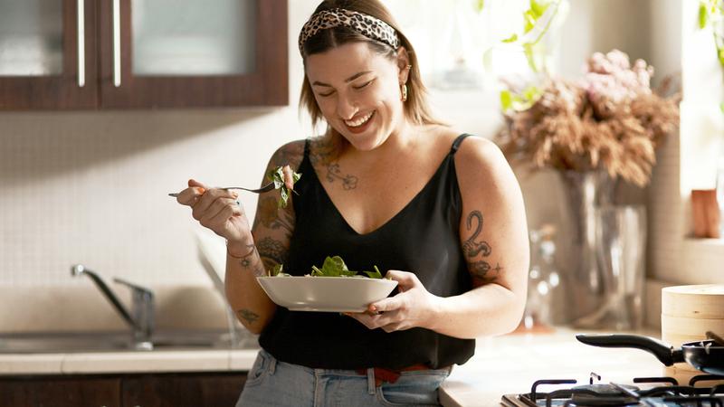 A young woman eats a bowl of salad while standing in her kitchen at home.