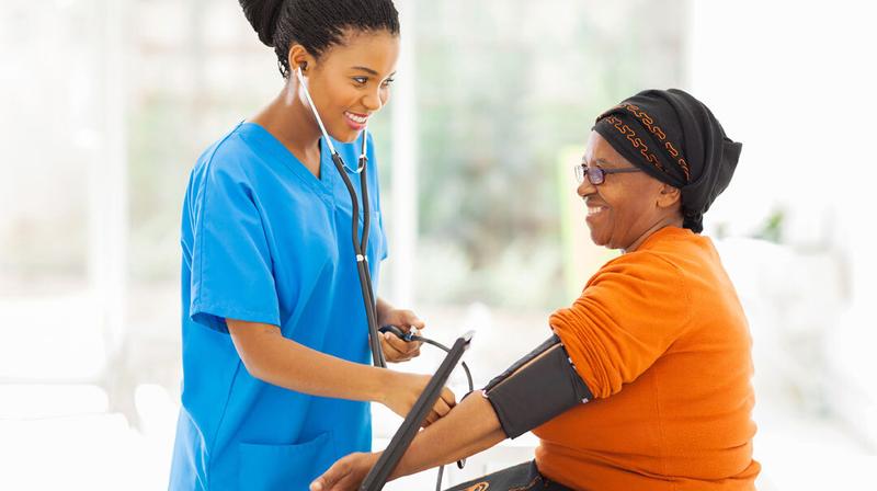 A healthcare provider wearing blue scrubs takes the blood pressure reading of a woman wearing a bright orange shirt and a head scarf.