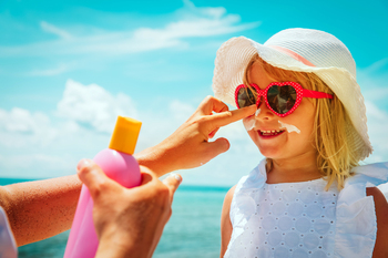 Close up photo of a little girl's face, wearing heart sunglasses, as her mother applies sunscreen lotion to her daughter's face.
