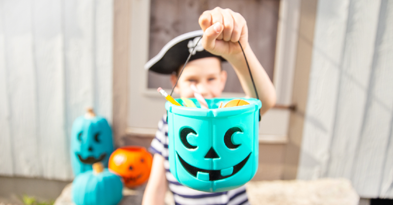 A child, wearing a pirate costume, holds a blue Halloween bucket full of candy.