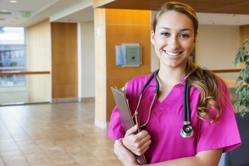 A young healthcare professional wearing pink scrubs, holds a clip board and poses for a photo in a hospital lobby.