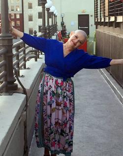 A woman with a shaved head and wearing a bright blue shirt and floral skirt smiles broadly as she stands on a balcony.