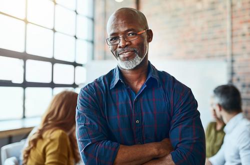 A middle-aged African American man stands in an office setting with his arms folded