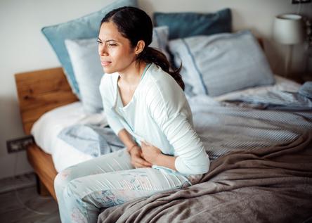 A young woman winces in pain while holding her stomach sitting in bed at home.