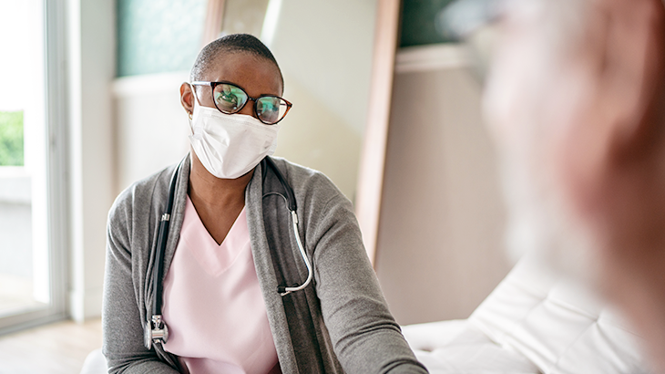 A nurse wearing a mask talks with a patient in a clinical setting.