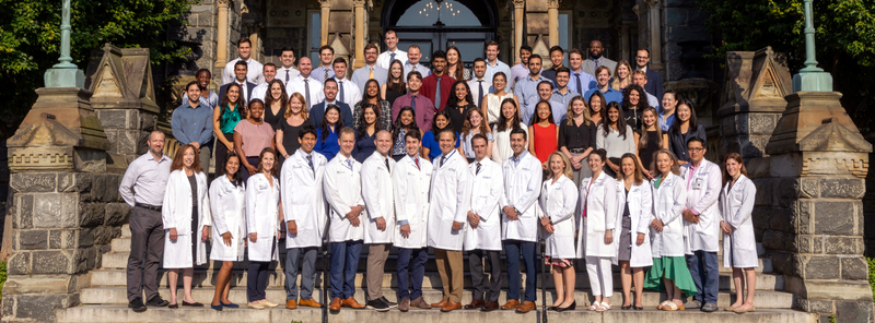 The current class of internal medicine residents at MedStar Georgetown University Hospital stands together on the steps of a historic stone building and pose for a class photo.