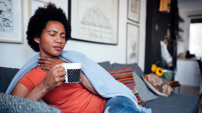 A woman sits on her sofa with a hot drink to soothe her sore throat.