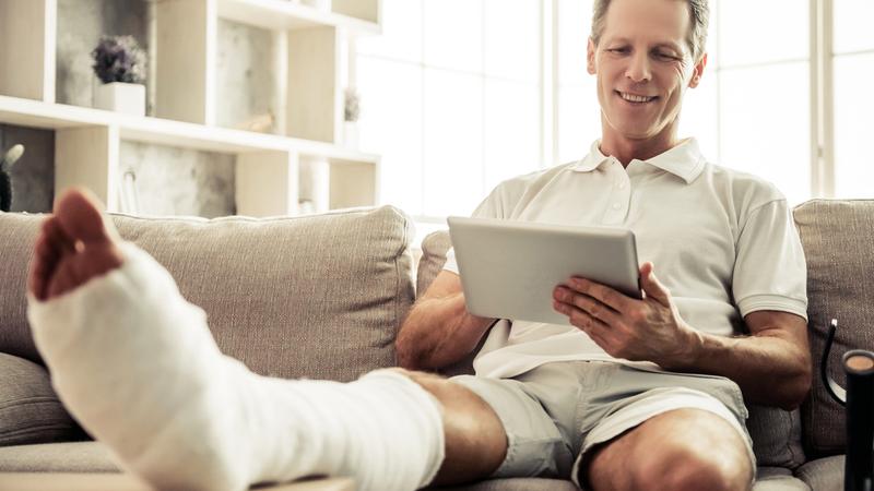 A man with a bandaged leg sits on the sofa in his home.