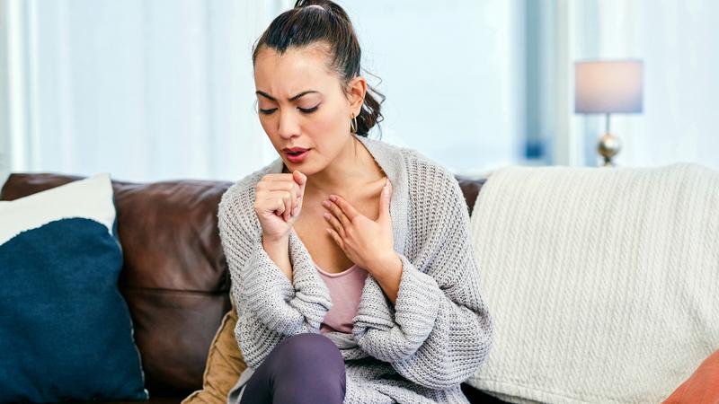 A young woman, feeling ill, is sitting on her living room sofa and coughing.