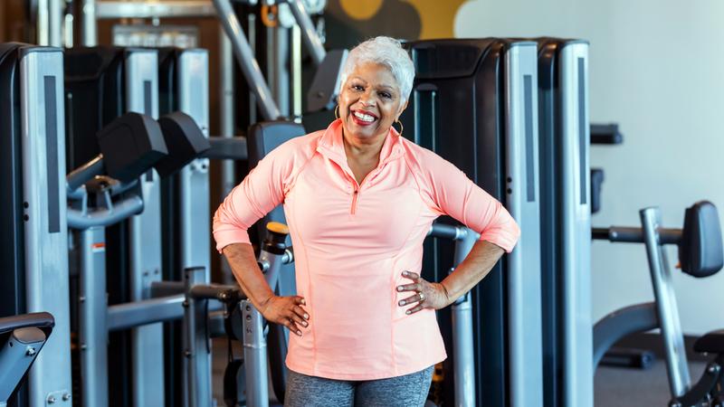 一位身穿运动服的老年妇女站在健身房的举重器械前，双手叉腰，对着镜头微笑。