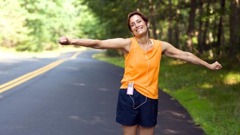 A female runner wearing an orange shirt stretches her arms out to the side and smiles for the camera.