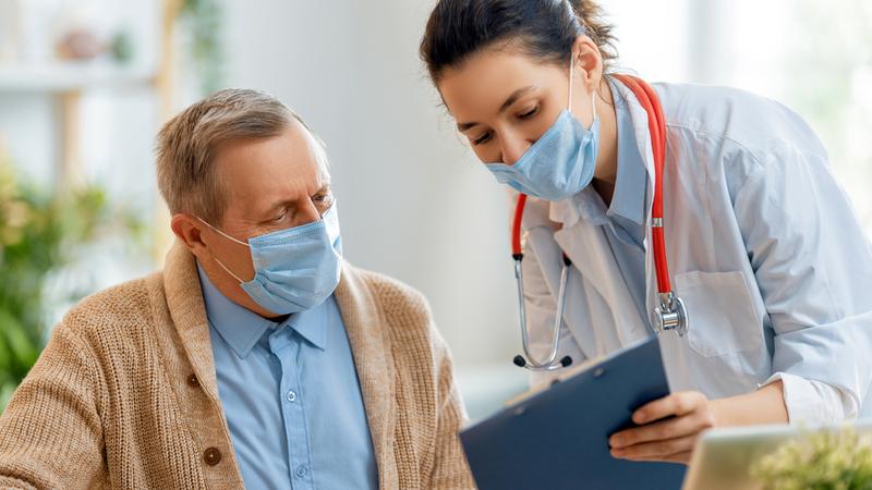 A young female medical professional holds a clip board and talks to an older male patient. Both people are wearing masks.