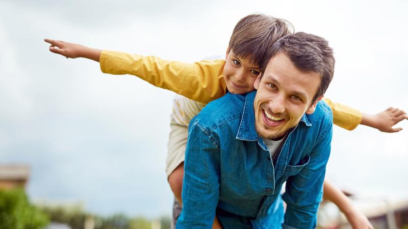 A man holds a boy on his back and the boy has his arms outstretched as if playing airplane. They are outside in a park-like setting.