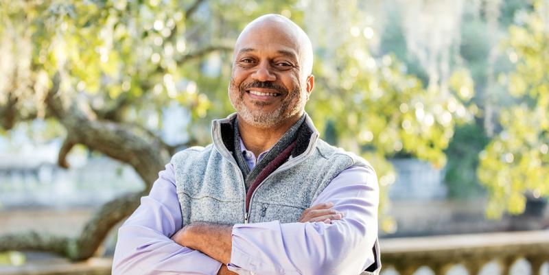 Headshot of a mature African-American man, in his 50s, taking a walk in the park. He is wearing a warm vest and long sleeves for the cool weather. He is smiling at the camera with arms crossed.