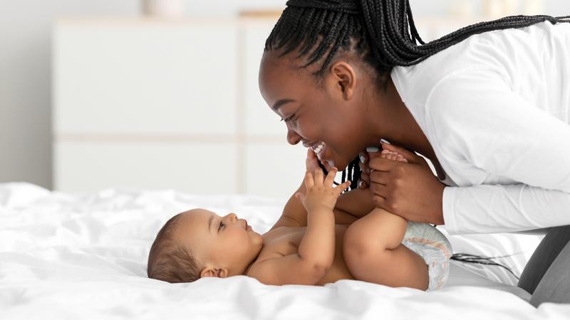 An african-american woman smiles at her infant who is laying on a bed at home.