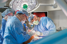 Dr Ezekiel Molina wears full operating protection and stands for a portrait in an operating room at MedStar Washington Hospital center. He is wearing a mask, gown, face shield, a surgical skull cap and magnifying headset.