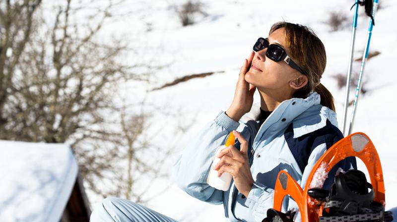 A woman, on a ski slope, puts sunscreen on her face.