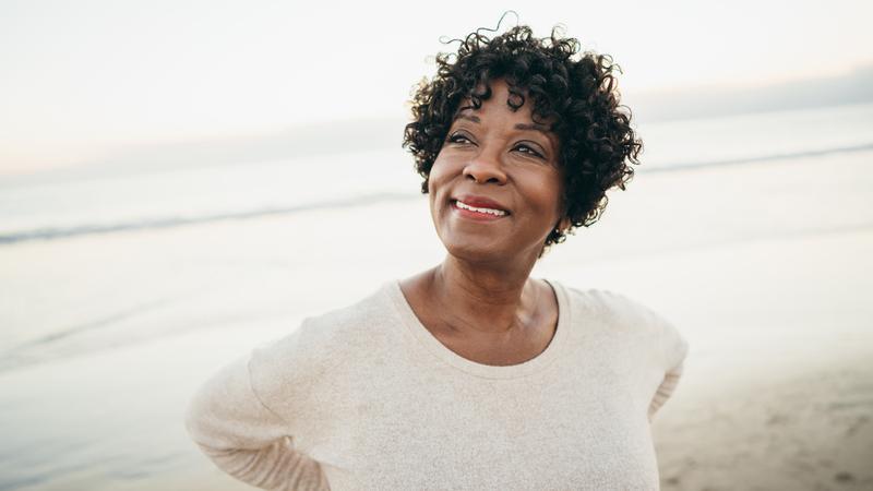 An african-american woman stands on a beach and smiles as she looks off into the distance.