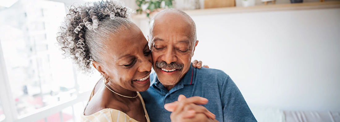 A mature couple smiles as they dance cheek-to-cheek at home.