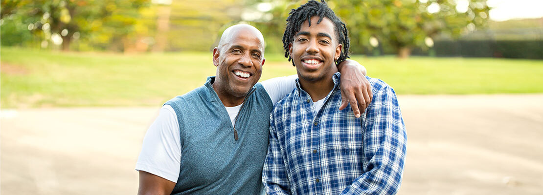 An older man stands with his arm around the shoulder of a younger man as they both smile for the camera.