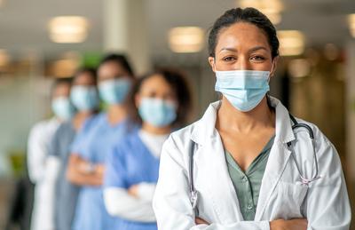 A group of healthcare professionals, wearing masks, stands in a line to pose for a photo in a hospital hallway. All are wearing masks. The people in the back of the line are out of focus.