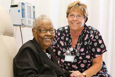 A nurse sits with a patient in an infusion center. They are both looking at the camera and smiling.