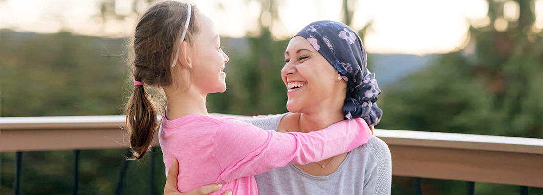 A young mother fighting cancer and wearing a head scarf hugs her daughter and smiles at her deeply as they share a few moments of tranquility together outdoors on a deck.