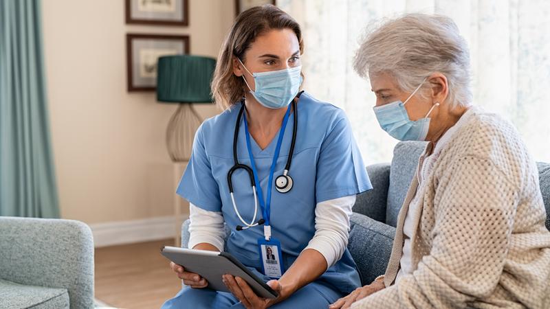 Young nurse and senior woman talks to a senior woman during home visit and wearing face mask. The nurse is holding an ipad. Both women are wearing masks.