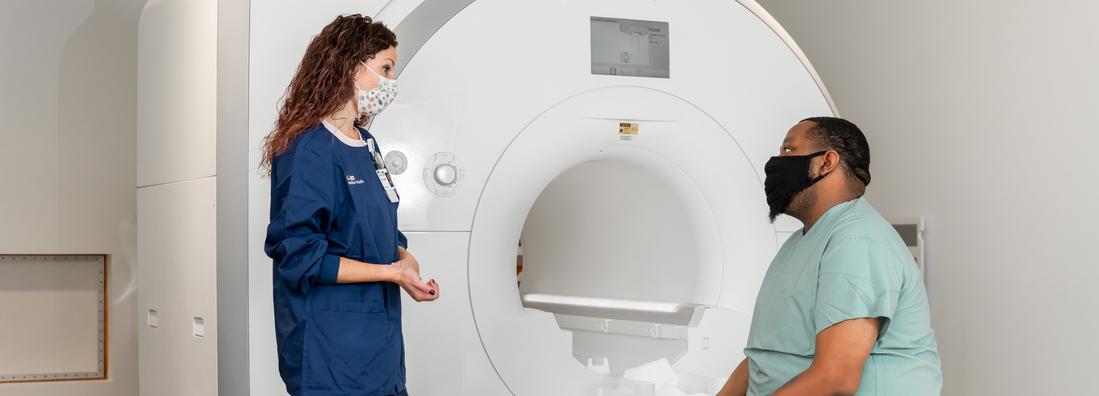 A radiology technicians talks with a patient who is seated on the table of a radiology scanning machine. Both people are wearing masks.