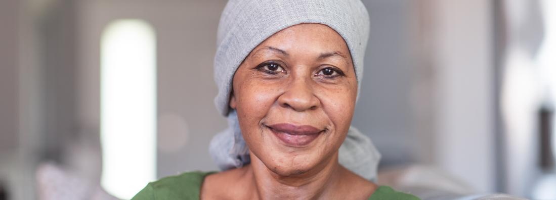 A mature black woman with cancer is wearing a scarf on her head. She is sitting at home in her living room. She is smiling directly at the camera.