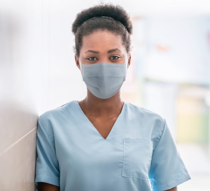 A nurse, wearing a face mask, leans against a wall and looks at the camera.
