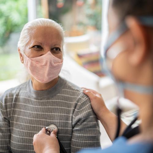 Nurse listening to senior patient's heartbeat during home visit. Both women are wearing masks.