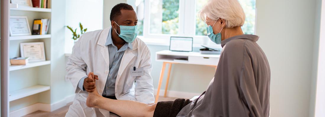 A doctor examines the leg of a senior female patient during an office visit appointment. Both people are wearing masks.