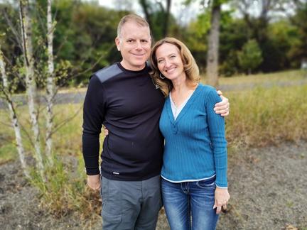 Mike Isreal stands with his arm around his wife as they stand outdoors on a wooded path..