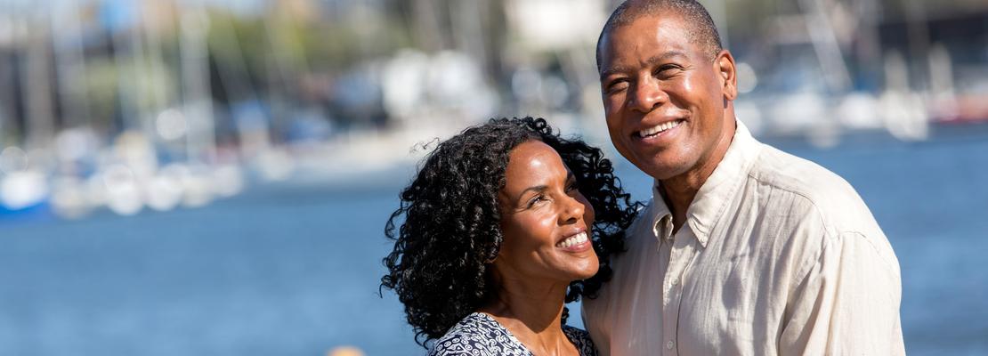 A mature african american couple hold hands near the water with a harbor in the background.