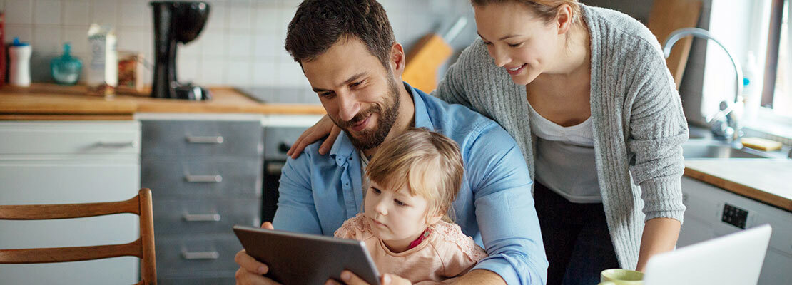 A family with a young child looks at an ipad while at home in their kitchen.