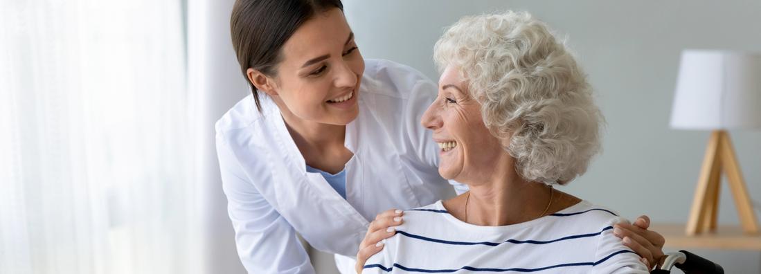 Smiling elderly woman sits in wheelchair and talks with a caring young nurse in living room.