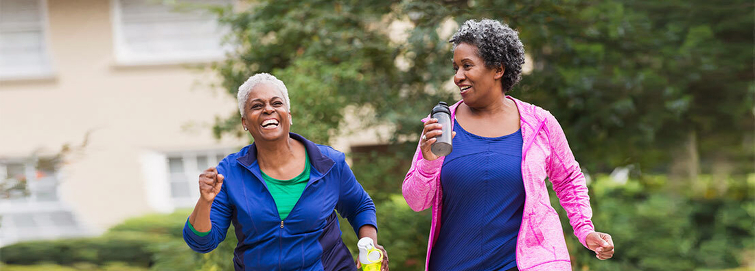 Two senior African American women getting in shape together. They are jogging or power walking on a sidewalk in a residential neighborhood, talking and laughing.